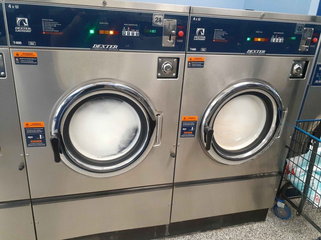 Thick white suds fill the round glass doors of two stainless steel Dexter front-load washers mid-cycle, showing what happens when too much detergent goes into a laundromat washer in Hemet, CA.