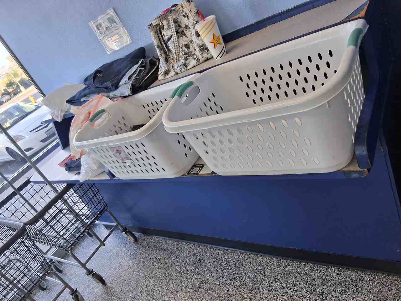 Laundry Baskets on Folding Counter San Jacinto Laundromat-1280 Two white plastic laundry baskets with teal handles sitting on a navy blue folding counter. Folded clothes and bags are behind them, wire laundry carts are in front, and a parked car shows through the window, inside The Laundry Hamper in San Jacinto, CA.