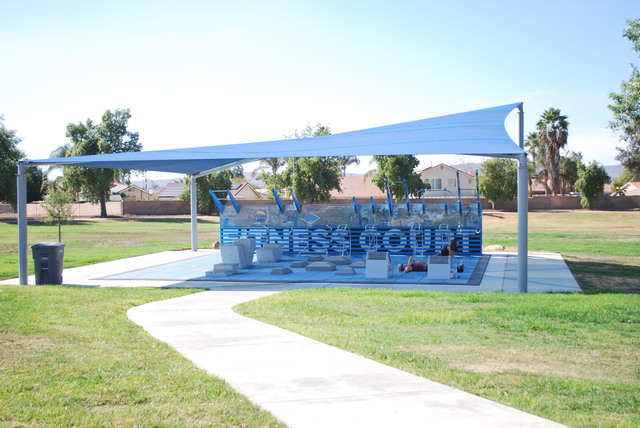 Outdoor Fitness Court at Mary Henley Park, with a blue shade sail canopy over a blue exercise zone with workout stations and a curved concrete path across the grass — a neighborhood park spot near The Laundry Basket in Hemet, CA.