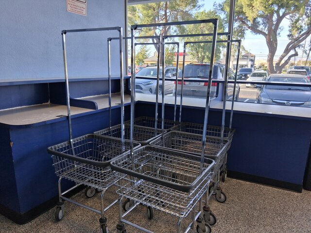 Rolling Laundry Carts by Folding Counter Laundry Basket Hemet CA-640 Four chrome rolling laundry carts with wire baskets and hanging racks sit beside a gray folding counter with navy blue trim, in front of a large window that looks out on the parking lot at The Laundry Basket in Hemet, CA.