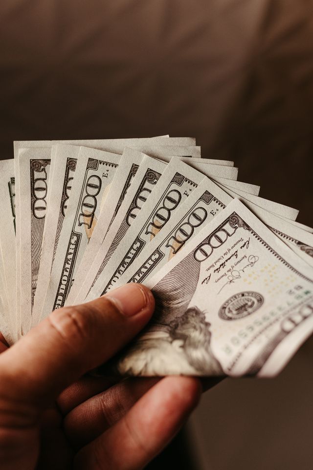 hand-with-dollars (image by: jonathanborba of Pexels.com) fanned out display of $100 dollar notes held in a person's hand on a desk.